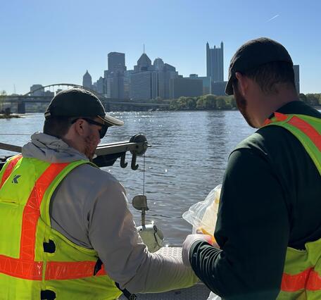 Two people on a boat collecting water samples with downtown Pittsburgh skyline in the background