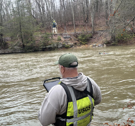 Two USGS hydrologists stand on either side of a river working on ADCP measurement and OTJ cableway training