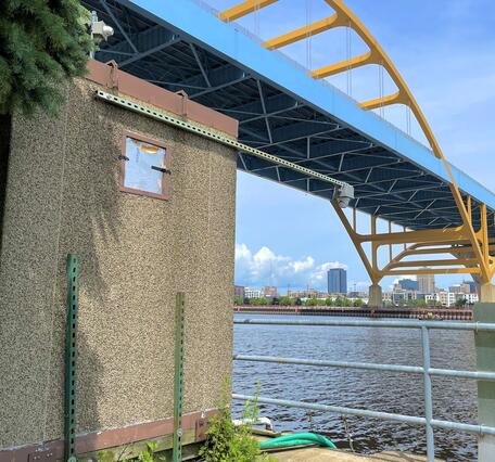 Streamgage housing next to a river with a large suspension bridge in the background