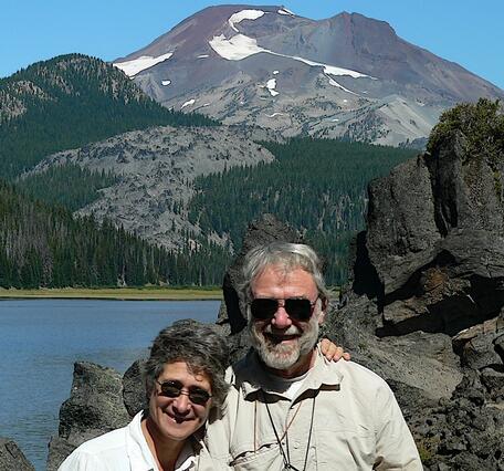 An older man and woman standing in front of a rock outcrop with a lake and forested volcano in the background. Both are wearing khaki shirts, sunglasses, and hand lenses. 