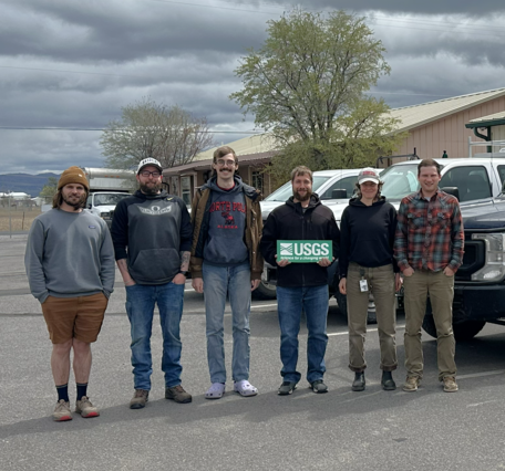 Six people stand in a row in a parking lot with trucks outside single story rectangular buildings on a cloudy day
