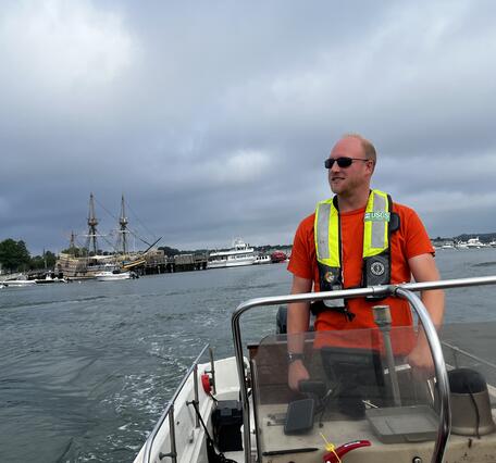 Man with life preserver driving boat with large ship in the background.