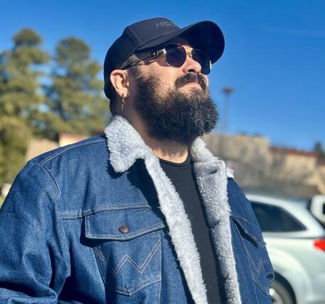 Kelvin, a bearded man with sunglasses, a black cap, and a denim jacket, looking up into the sky
