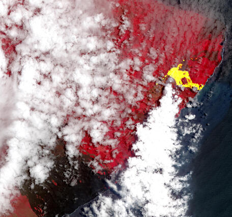 Satellite image with a lot of clouds plus an erupting volcano on an island where the background vegetation is depicted in red