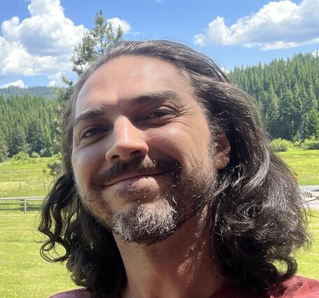 An image of Russell Krantz smiling in front of conifer trees and a green field