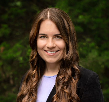A woman with long brown hair wearing a white shirt and a black blazer smiles for her picture.
