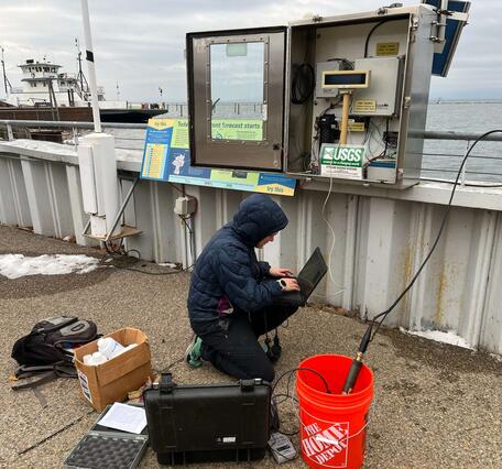 Hydrologic technician squatting down with a laptop next to a streamgage connected to a waterfront railing.