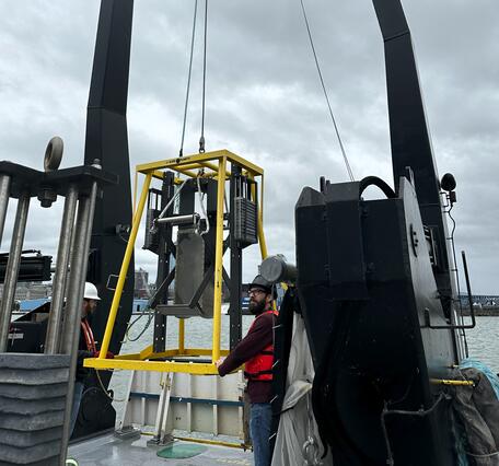 Two scientists guide sampling equipment attached to large crane, on to deck of research vessel