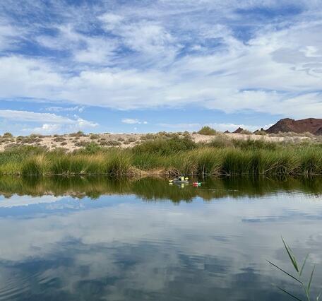 Person in yellow kayaking near far bank on Las Vegas Wash, towing stream gaging equipment under partly cloudy sky.