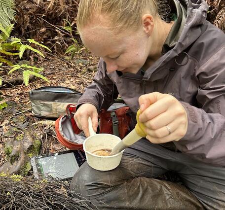 Person sitting on ground in rain gear holding a sample in a plastic cup and a baster.