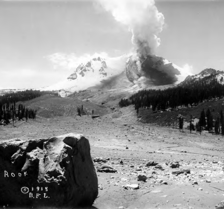 A black and white photograph looks across a swath cut through a pine forest by a debris and mud flow that originates at a steaming volcanic peak in the distance. A large crater is visible on the side of the volcano, which is emitting the steam. The rest of the volcano is heavily blanketed in snow.