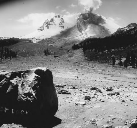 A black and white photograph looks across a swath cut through a pine forest by a debris and mud flow that originates at a steaming volcanic peak in the distance. A large crater is visible on the side of the volcano, which is emitting the steam. The rest of the volcano is heavily blanketed in snow.