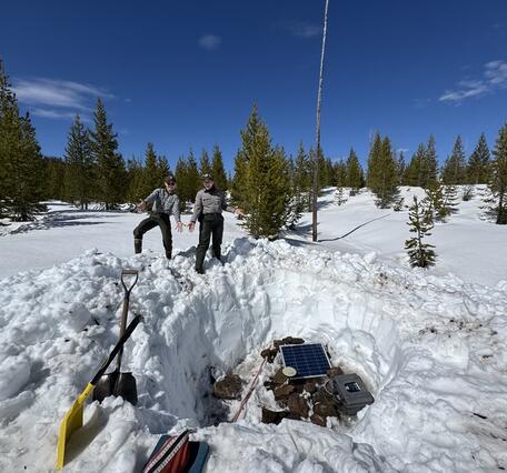 Two woman point out a pit in snowy landscape with equipment in the pit. Sparse trees and blue sky in background.