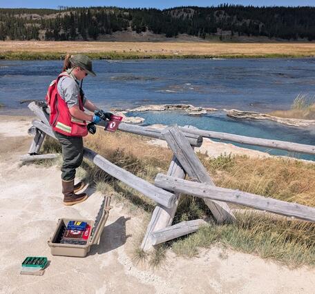 Woman in red vest fastens warning sign to wooden barrier. River, meadow, and forested hill in background.