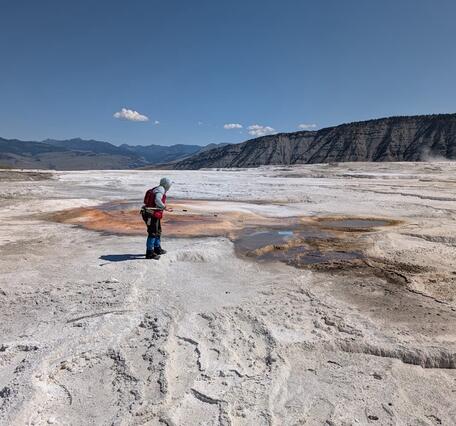 Woman in red vest standing on white terrace in front of reddish hot spring under blue sky. Mountains in the background.
