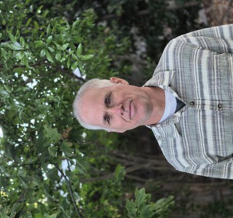 Smiling man in button up green and white striped shirt. Green trees form the back ground