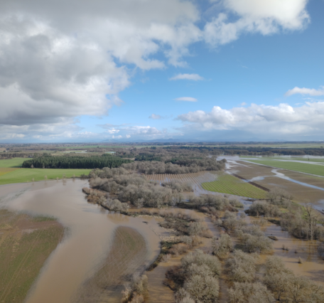 Sediment filled brown meandering river floods into nearby agricultural land. Partly cloudy day.