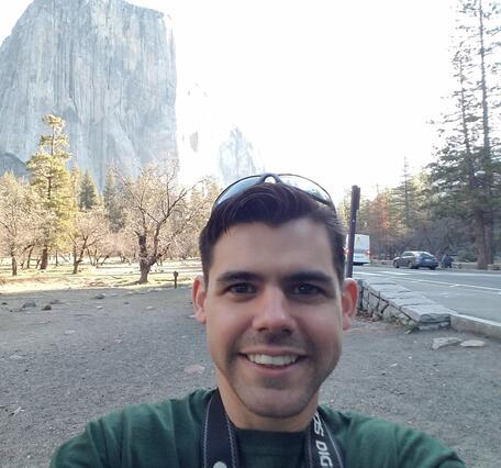 man takes selfie in front of Half Dome rock in Yosemite, trees and road in background
