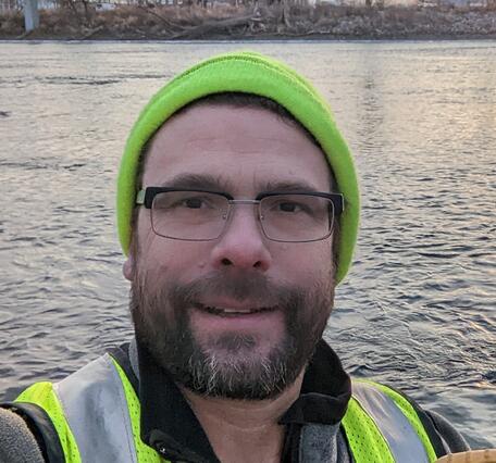 Man wearing glasses and bright green hat and vest taking selfie in front of river
