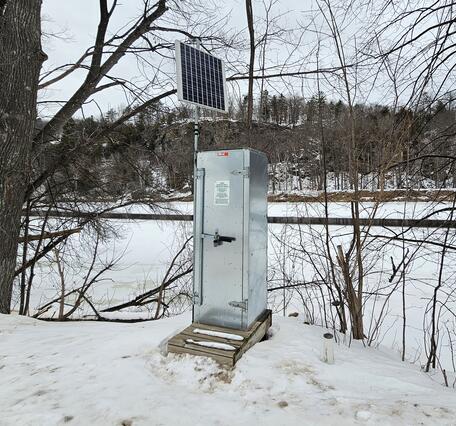 A gray metal streamgage housing with an attached solar panel sits on the shore of a snow-covered frozen river.
