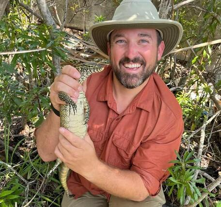 a man in a salmon-colored shirt and a tan hat holds a large black and white lizard, shrubs in the background