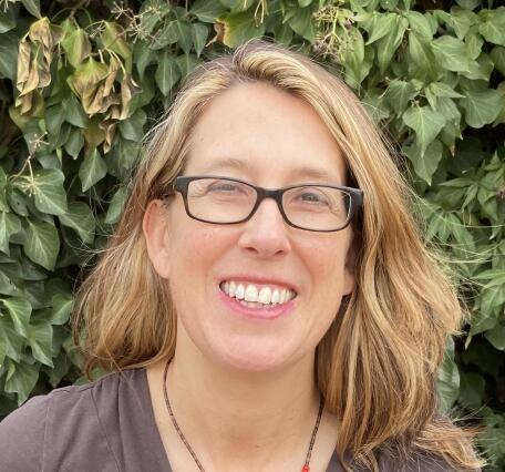 Headshot photo of a smiling woman with shoulder length blond hair and glasses, with vegetation in background