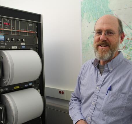 A man stands in front of a machine holding two drums covered in paper that records seismic data