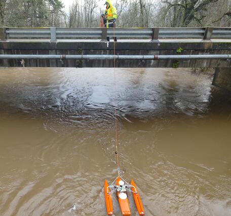 Female in safety gear pulls measurement equipment across the river from a bridge
