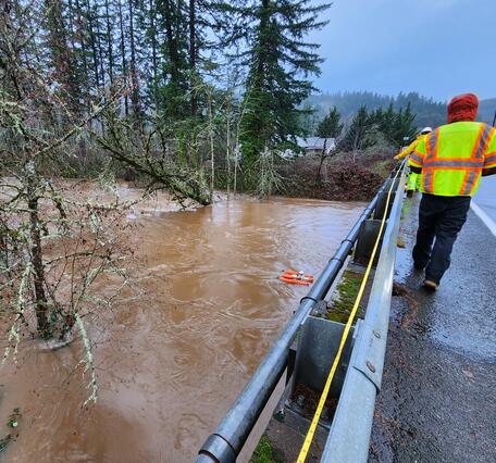 Swollen brown river water flowing through downed trees. Two technicians in safety gear pull ADCP across the channel 
