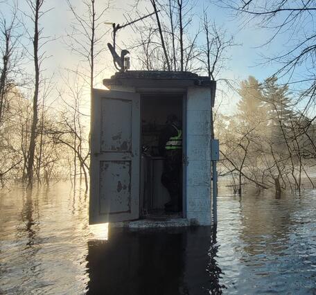 Hydrologic technician servicing a flooded streamgage housing, backlit by sunrise