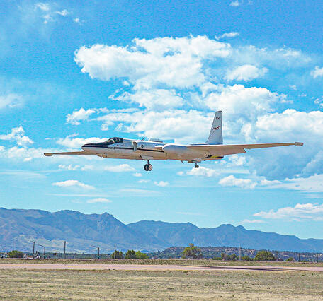 A white aircraft descends with landing gear extended set against a backdrop of blue sky, scattered clouds, and mountains