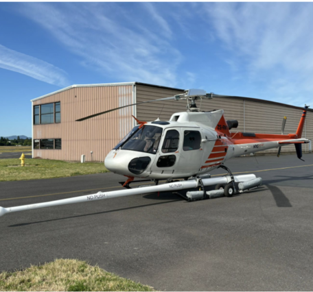 Red and white helicopter with long boom extending forward; helicopter is on the ground in front of building