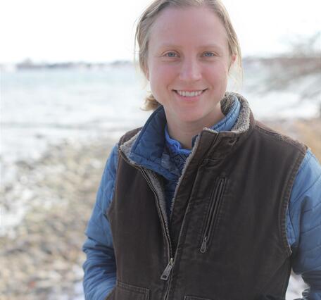 A woman with blonde hair in a long sleeved shirt and vest stands for her picture. 