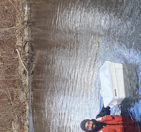 Man in a Lifejacket wading chest deep in a river with a styrofoam basket for samples