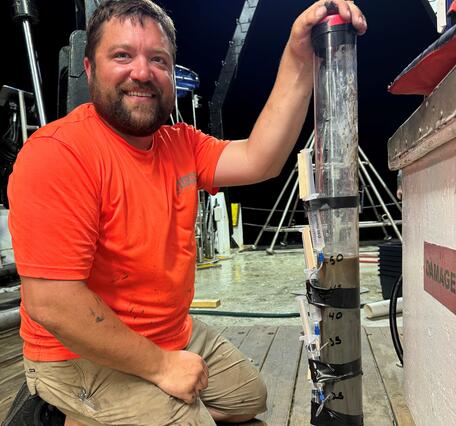 man kneeling on deck of ship next to a core sample at night