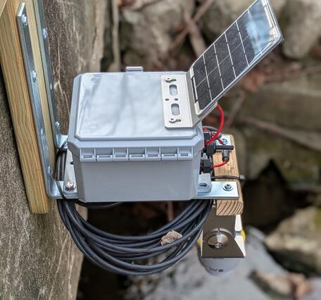 Close up view of gray box with solar panel affixed to bridge above stream