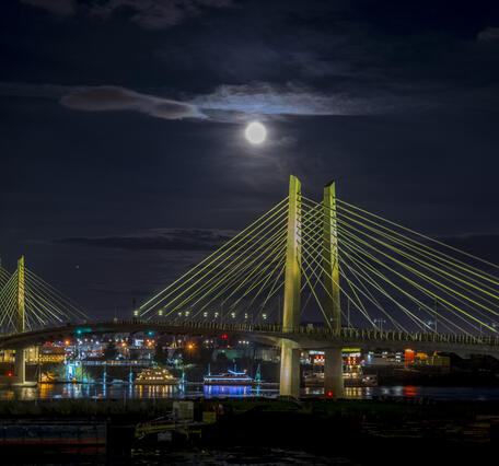 brightly lit colorful boats float below the bridge with its cables lit a pale yellowish green on a full moon night