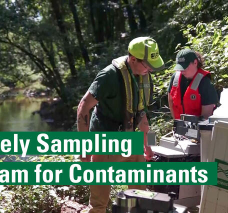 Two people lift heavy equipment adjacent to a stream. Text reads: Remotely Sampling A Stream for Contaminants.