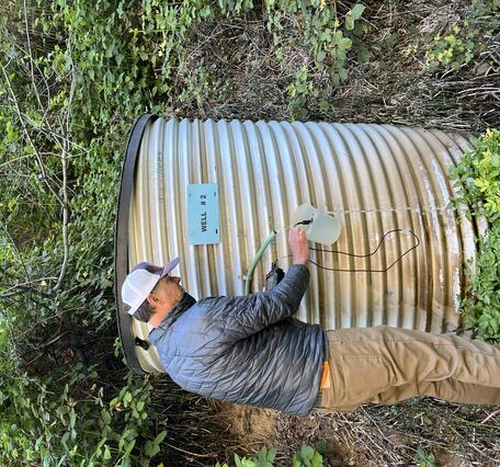 Tall man in a baseball cap uses a bucket to catch well water in to take measurements with a handheld sensor