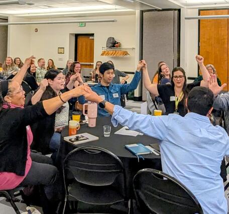 Groups of people at tables lift hands into the air to link up in unison