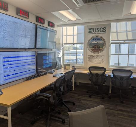 A small room holding a bank of large computer screens, multiple world clocks, and other communications equipment. A desk with multiple rolling chairs wraps around the perimeter of the room.