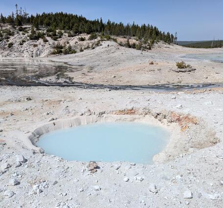 barren chalky ground with a milky-blue pool in the middle and a small hill with scrub vegetation in the background