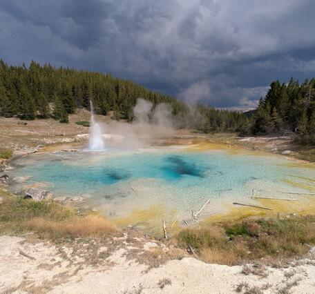 blue-water hot spring with a geyser to one side, forest in background, and storm clouds in the distance
