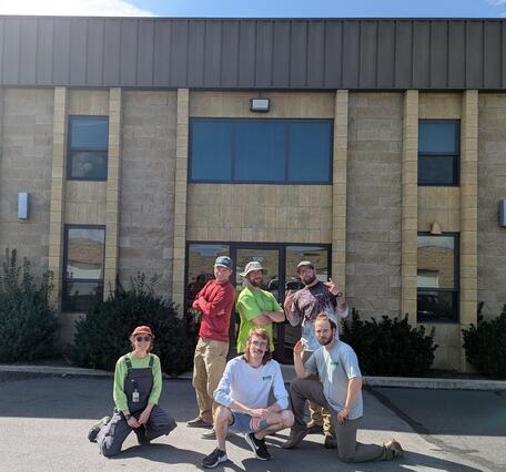 Six people pose in front of a brown brick building on a sunny summer day