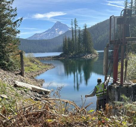 Man lowers a measuring tape into the lake's blue-green water. Island with trees reflect in the water. Mt. Hood in background.