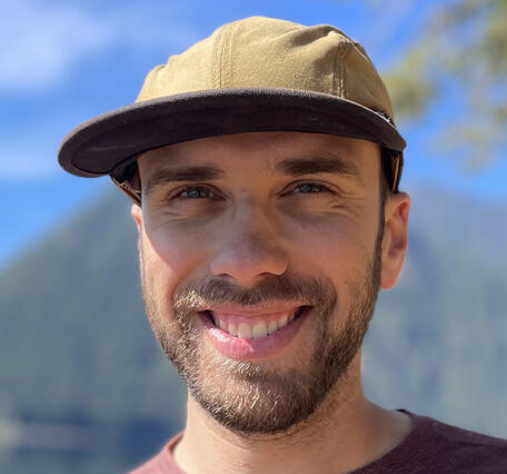 Photo of a young man wearing a fishing cap and smiling. He has blue eyes and brown hair and a beard. A mountain and lake are visible behind him.