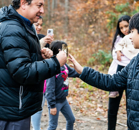 A man in a puffy black coat holds a small bird as a young boy reaches out to touch it