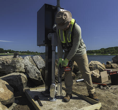 Person using drill to install anchor for gage monitor with river in the background.
