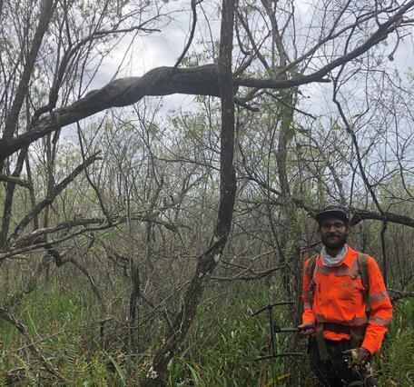 a man in bright orange shirt stands among grasses and bare trees, with a large python on a branch above