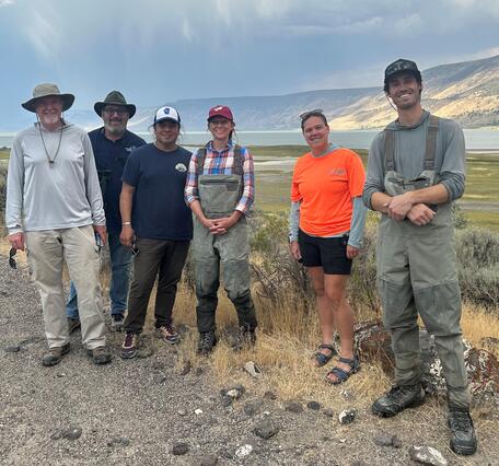 Five people in outdoor gear smile in front of a shallow lake on a sunny day